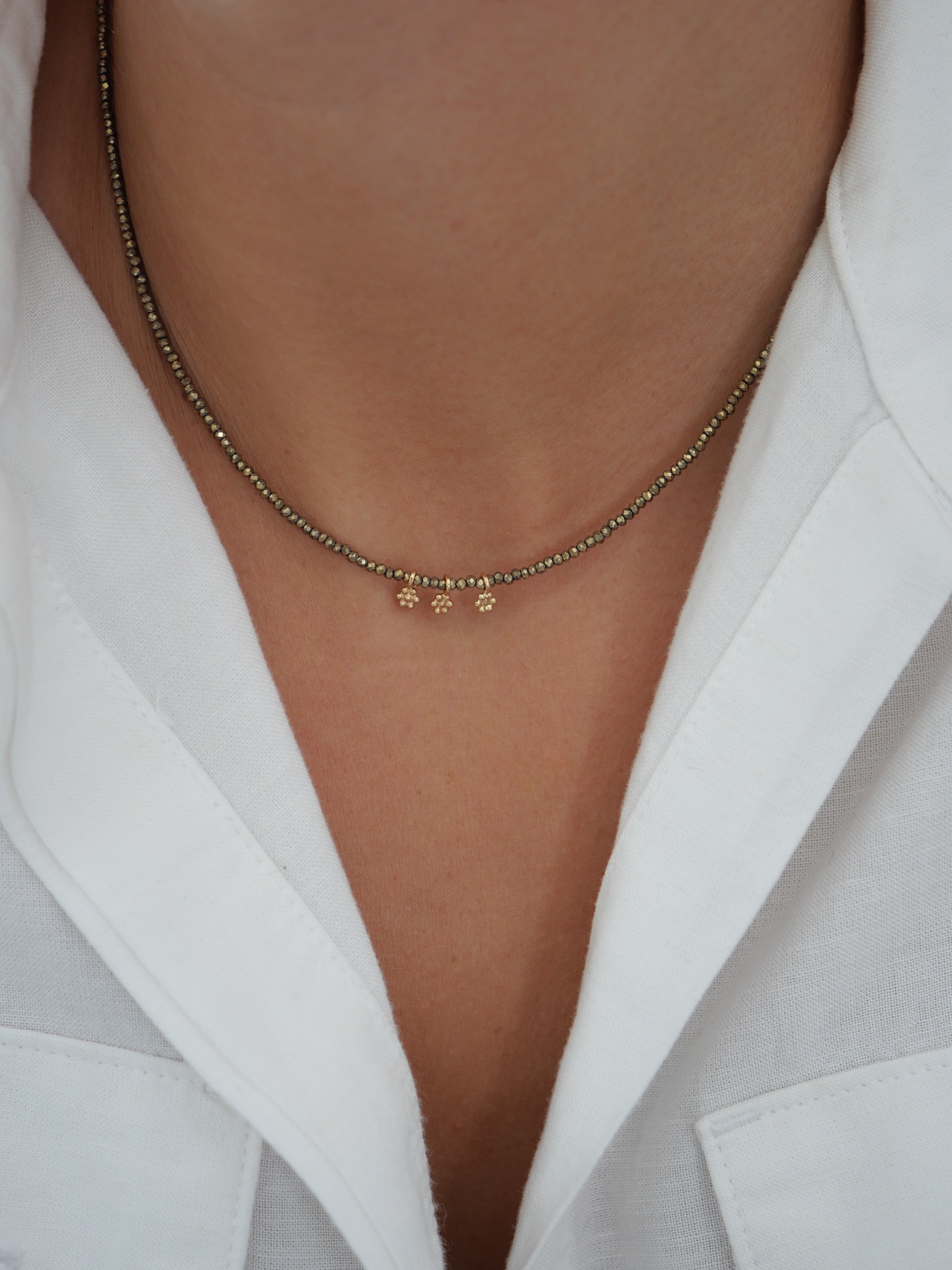 Collier Séléna 2 Collier ras de cou Séléna en hématite kaki facettée, orné de trois petites fleurs dorées, porté avec une chemise blanche entrouverte.