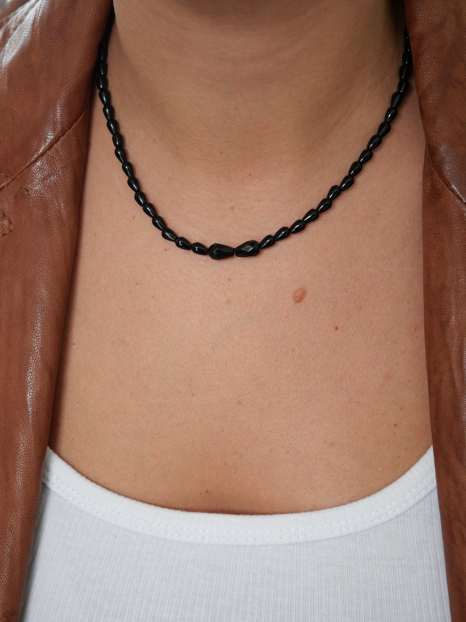 Collier Sweet rebel 1 Collier Sweet Rebel en onyx noir porté avec un débardeur blanc et une veste en cuir marron, détail des deux pierres centrales facettées.