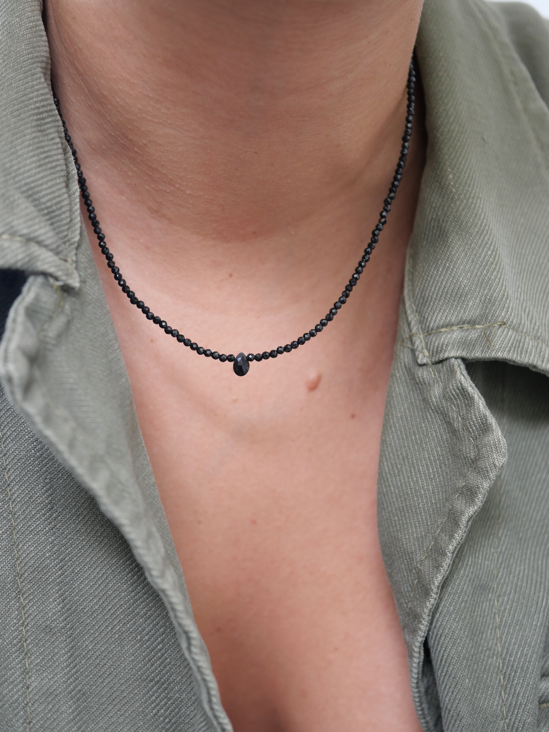 Collier Skinny Love 1 Collier Skinny Love en fines perles d’onyx noir facettées et goutte centrale, porté dans l’ouverture d’une chemise kaki.