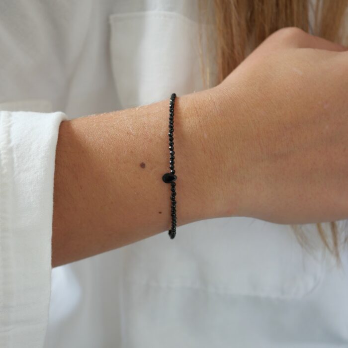 Bracelet Skinny Love en fines perles d’onyx noir facettées avec une petite goutte centrale, porté au poignet avec une chemise blanche.
