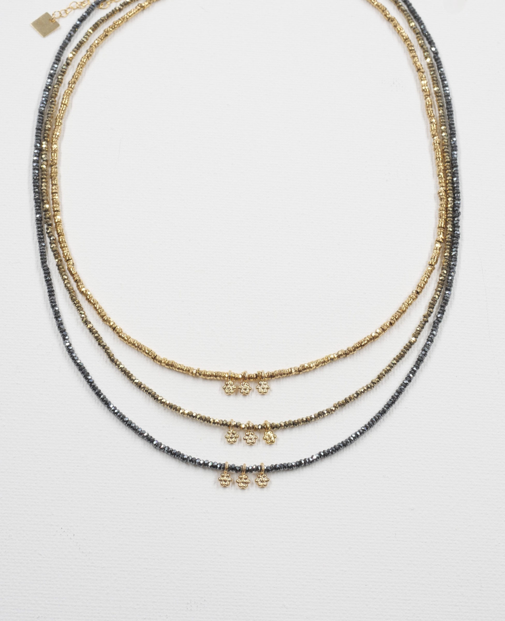 Collier Séléna 1 Trois colliers ras de cou Séléna en hématite facettée dorée, kaki et grise, chacun orné de trois petites fleurs dorées, présentés sur fond blanc.