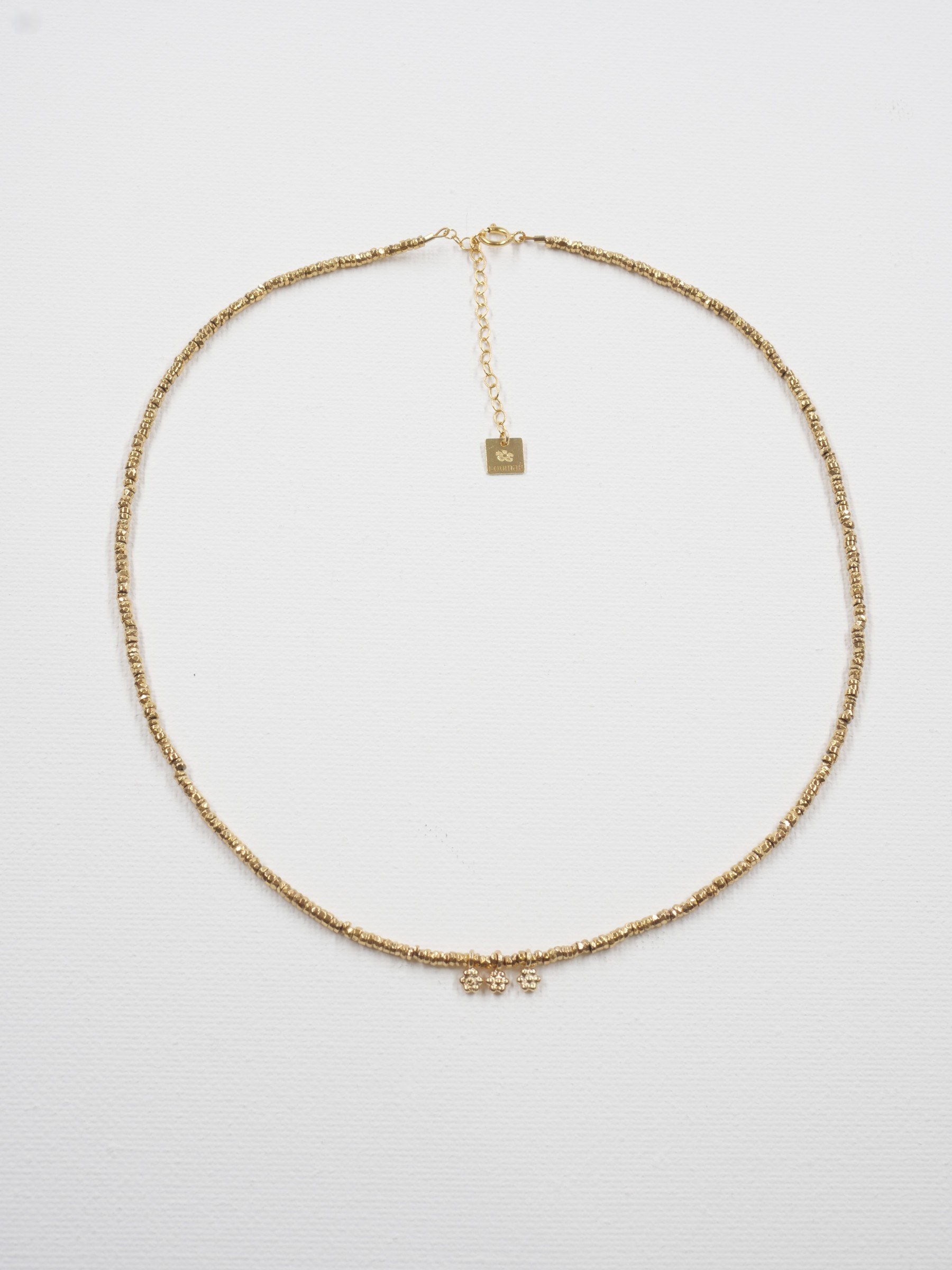 Collier Séléna 4 Collier ras de cou Séléna en hématite dorée facettée, avec trois petites fleurs dorées, fermoir et chaîne de rallonge, présenté à plat sur fond blanc.