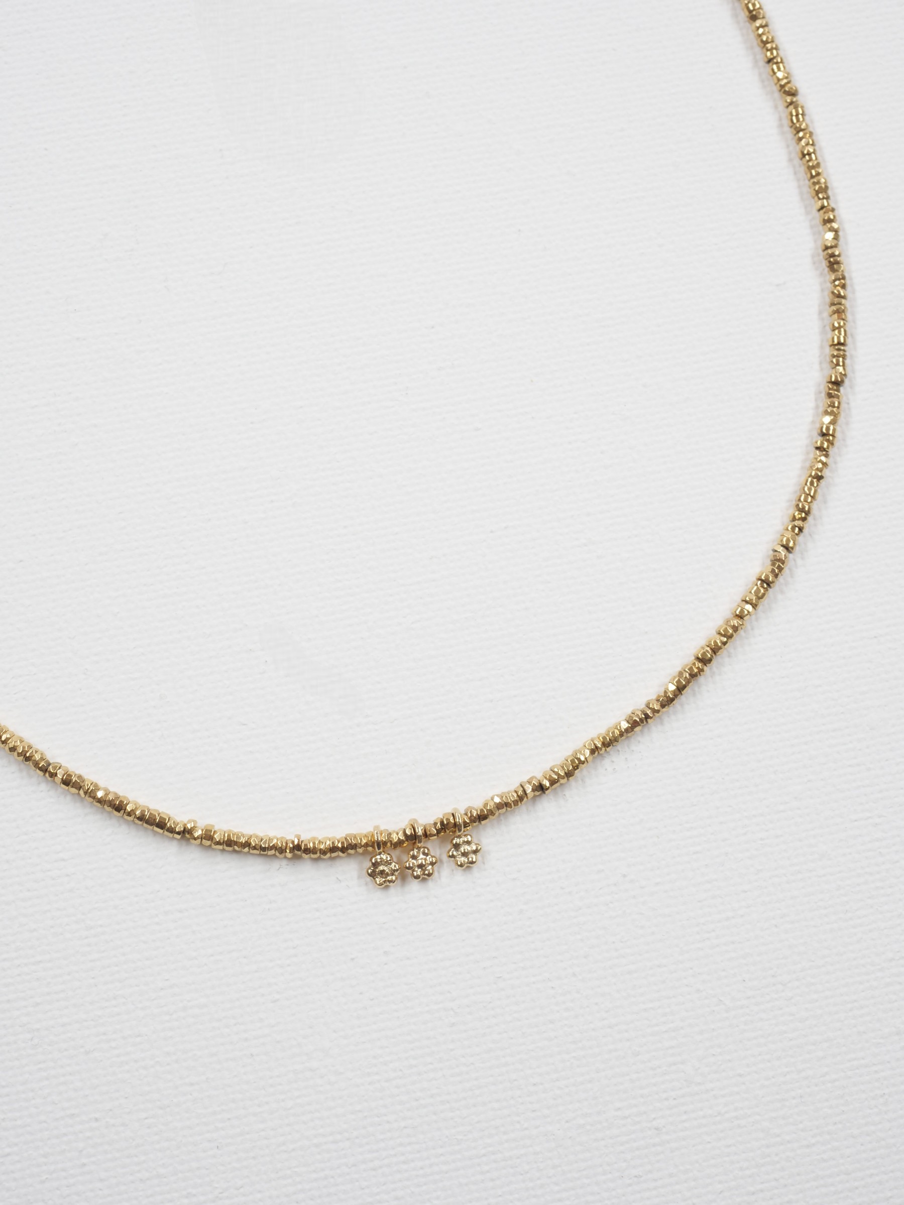 Collier Séléna 5 Collier ras de cou Séléna en hématite dorée facettée de 2 mm, orné de trois petites fleurs en laiton plaqué or, sur fond blanc.