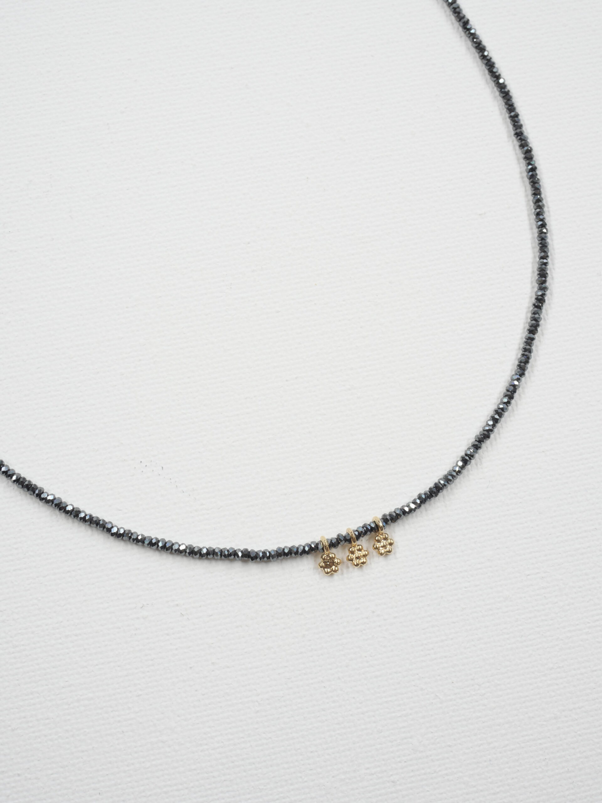 Collier Séléna 7 Collier ras de cou Séléna en hématite grise facettée de 2 mm, orné de trois petites fleurs en laiton plaqué or, sur fond blanc.