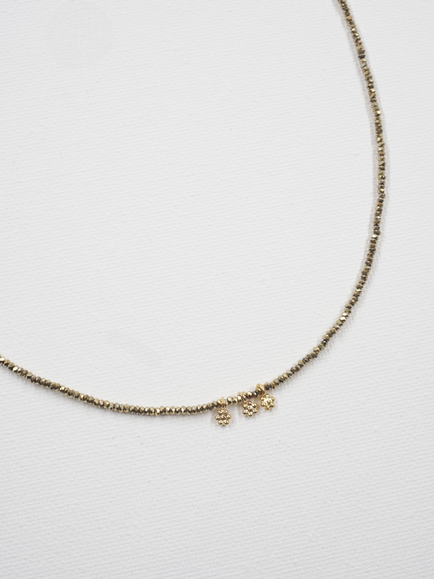 Collier Séléna 6 Collier ras de cou Séléna en hématite kaki facettée de 2 mm, orné de trois petites fleurs en laiton plaqué or, sur fond blanc.