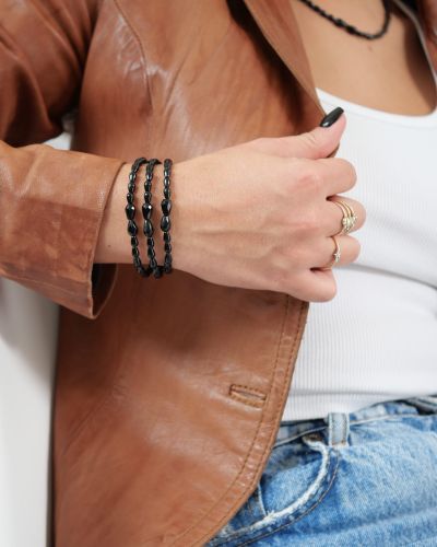 Trois bracelets Sweet Rebel en onyx noir portés en accumulation au poignet, avec pierres centrales facettées et veste en cuir marron.