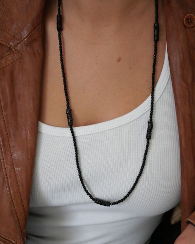 Sautoir Black Betty en perles et rondelles d’onyx noir facettées, porté sur un débardeur blanc avec une veste en cuir cognac.