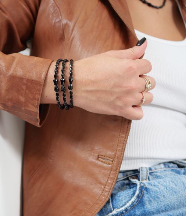 Trois bracelets Sweet Rebel en onyx noir portés en accumulation au poignet, avec pierres centrales facettées et veste en cuir marron.