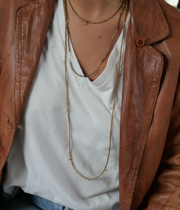 Collier Célia 5 Sautoir Séléna doré en hématite et petites fleurs plaquées or, porté en accumulation sur un tee-shirt blanc avec une veste en cuir.